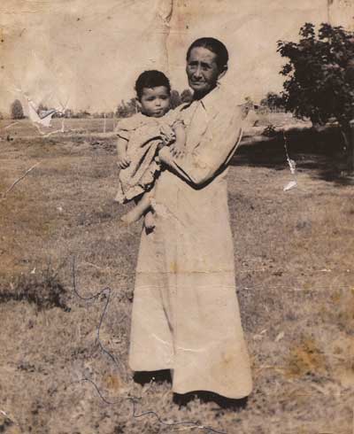 Clara Martínez holding her grandson, Robert Marron Romero, with a grassy field and a few trees behind. Links to full image.